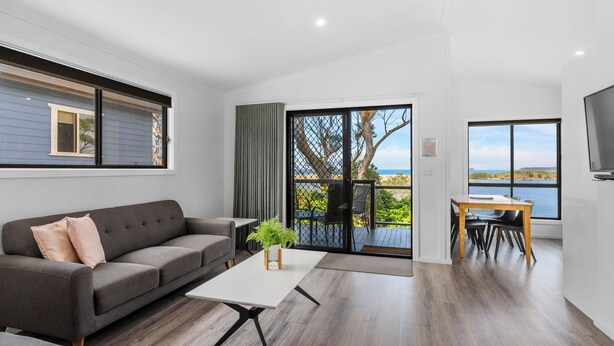 Un salon moderne avec un canapé gris, une table basse et un coin repas comprenant une table et des chaises. Une grande fenêtre offre une vue sur la mer.