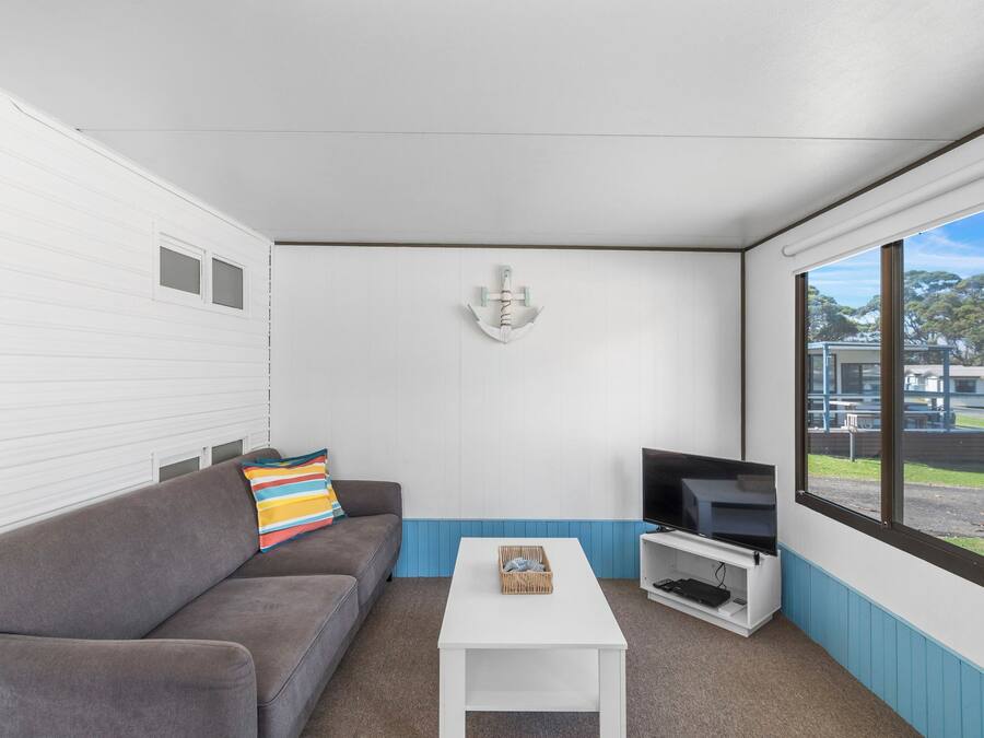 Una habitación con un sofá gris, una mesa de centro blanca, un televisor de pantalla plana y un ventanal con vistas al exterior.