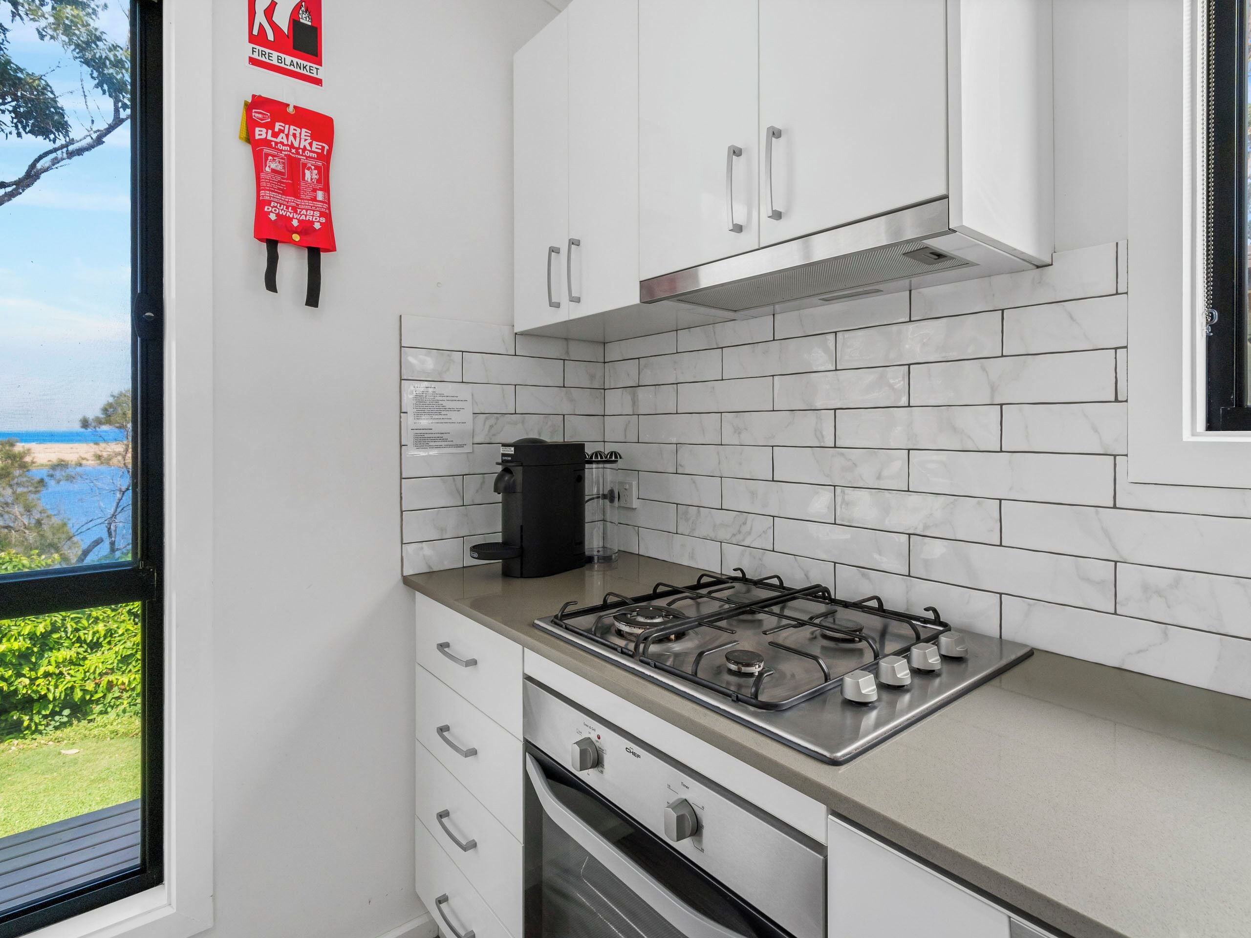 A modern kitchen with white cabinets, a gas stove, and a coffee maker. There is a window with a view of the beach.