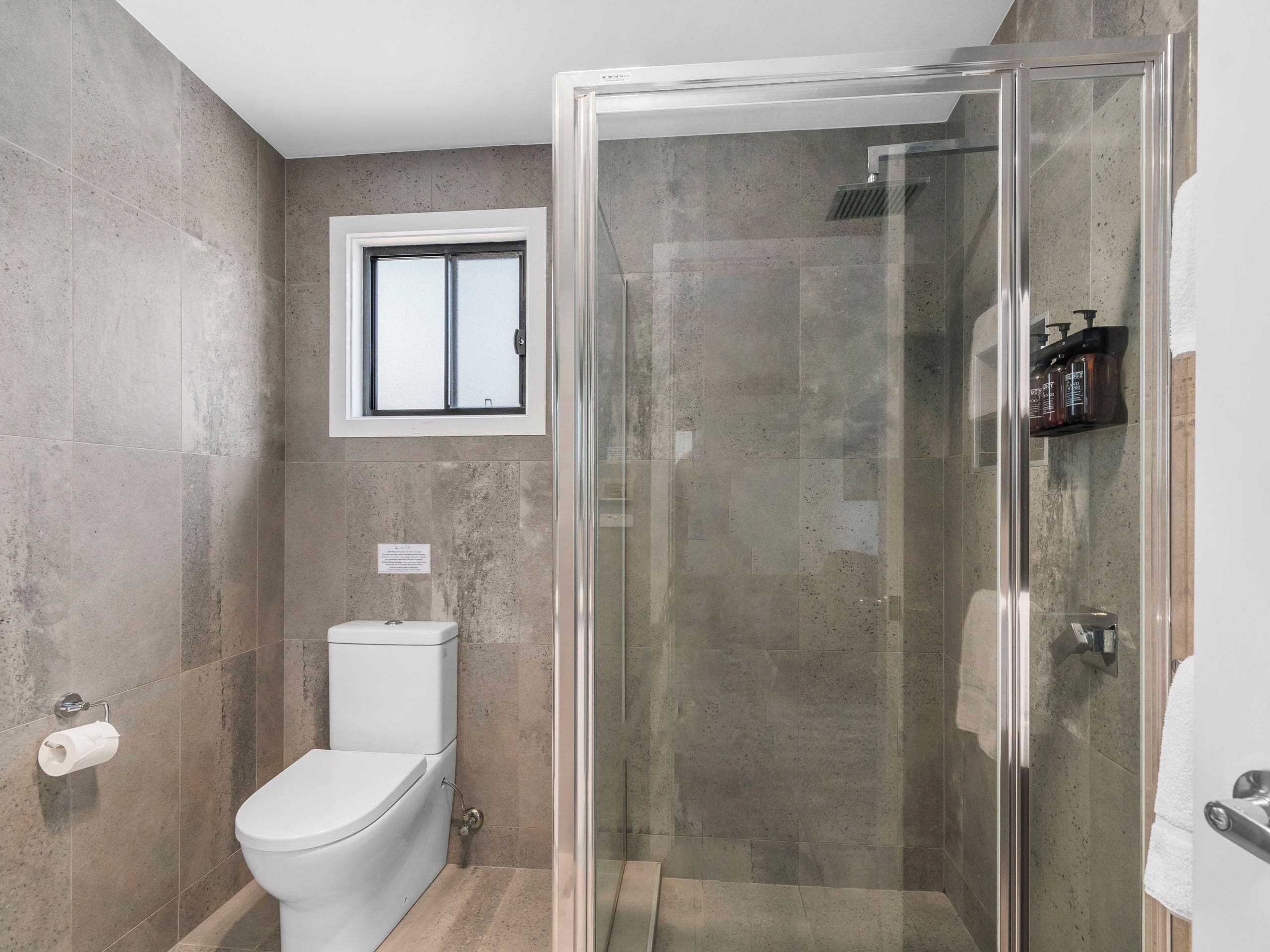A modern bathroom with a glass-enclosed shower, a toilet, and a window above the toilet.