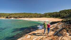 Un couple se promène sur une plage rocheuse, l’eau est claire et, en arrière-plan, on aperçoit une forêt.