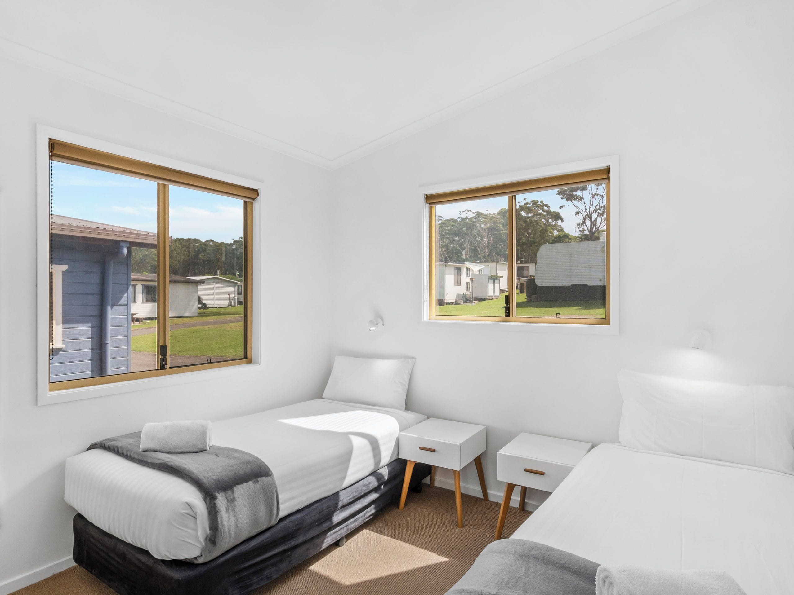 A modern bedroom with a bed, bedside tables, and a desk. There are two windows providing views of the outside.