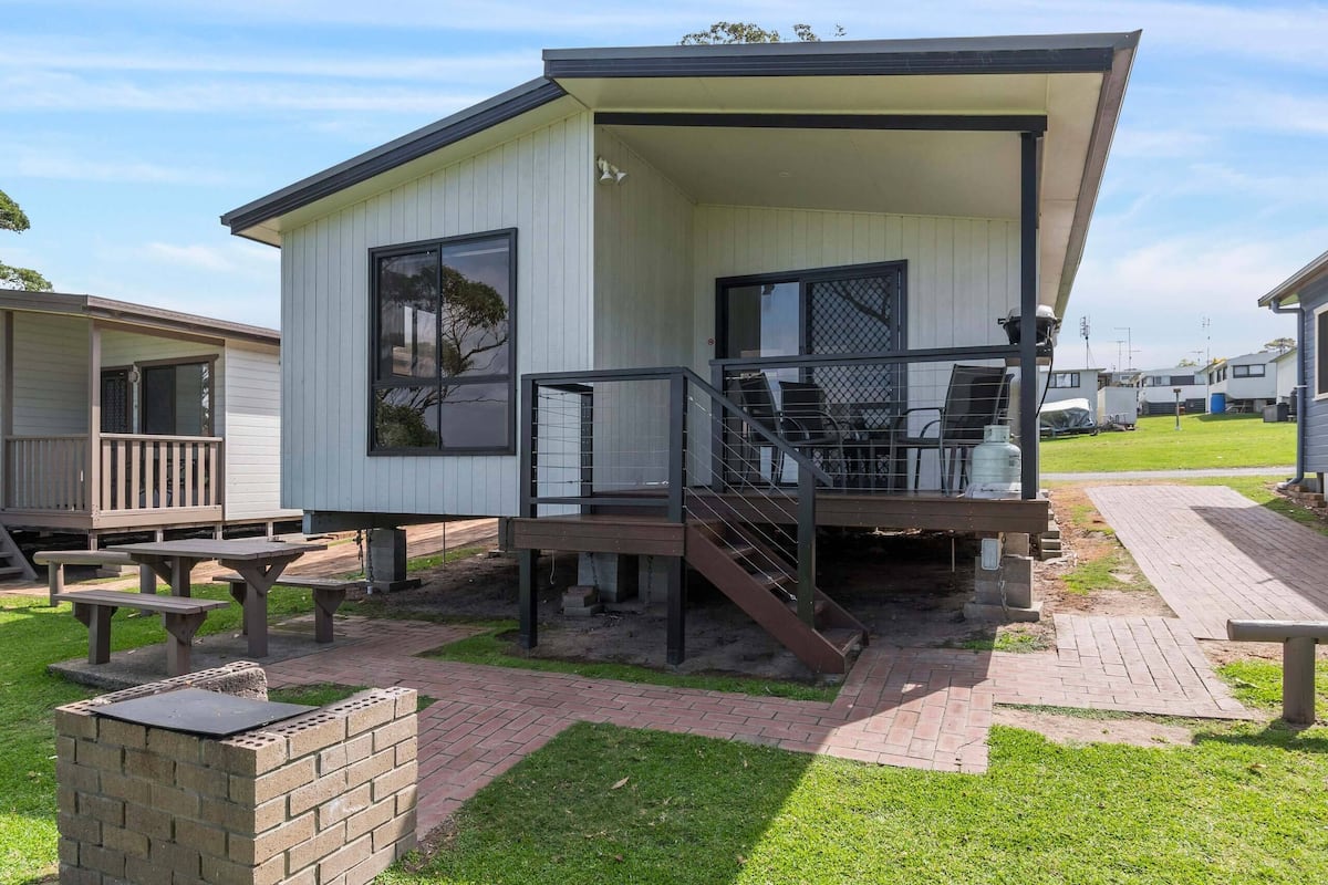 A single-story mobile home with a covered porch, a brick barbecue area, and a small outdoor seating space.
