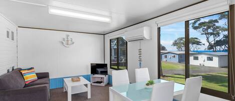 Una habitación con una decoración en blanco y azul, un sofá gris, una mesa de comedor blanca con sillas, un televisor de pantalla plana y un amplio ventanal con vistas al exterior.