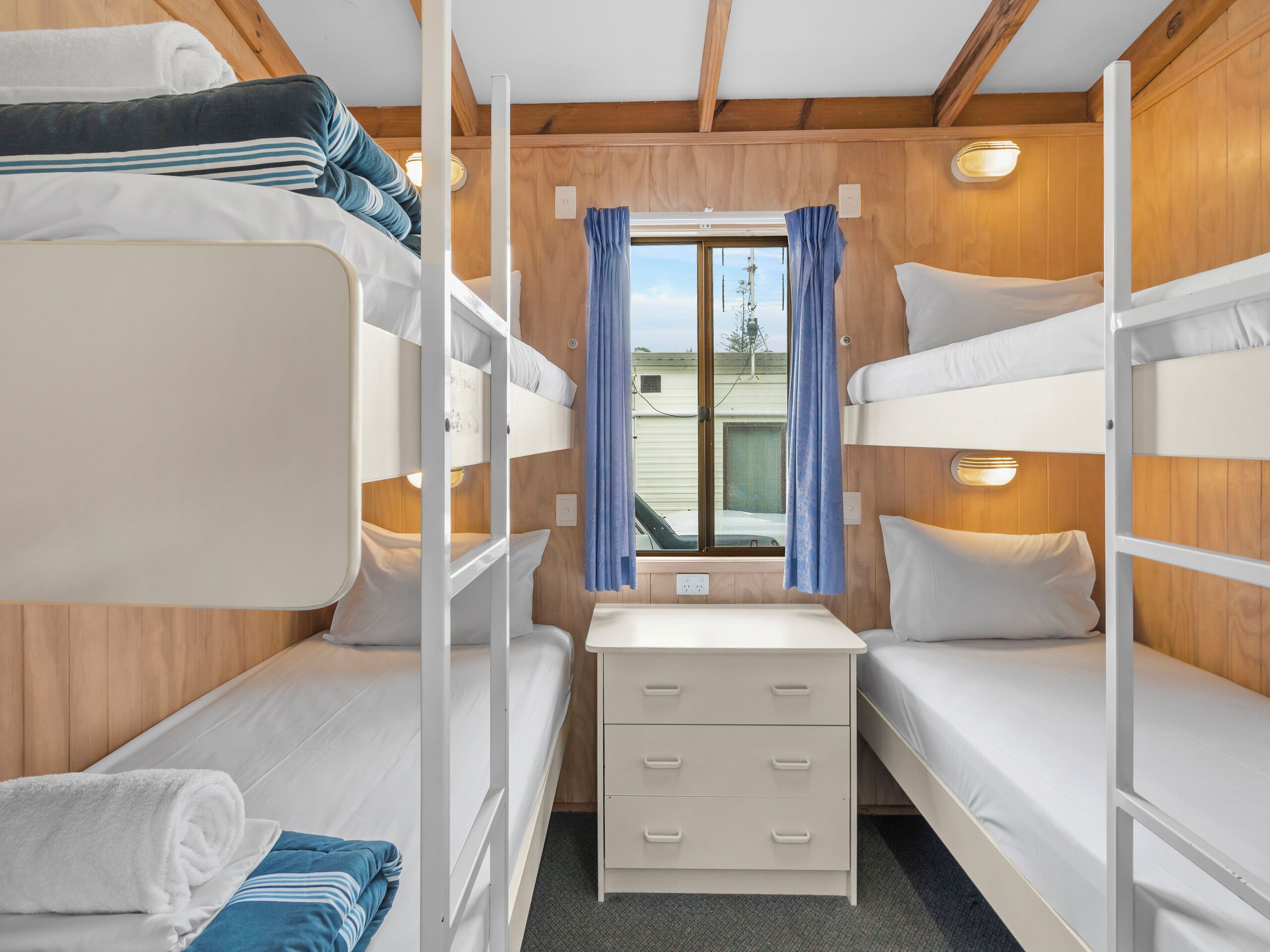 Une chambre avec des lits superposés, comprenant trois lits, une petite table avec des tiroirs et une fenêtre avec des rideaux.