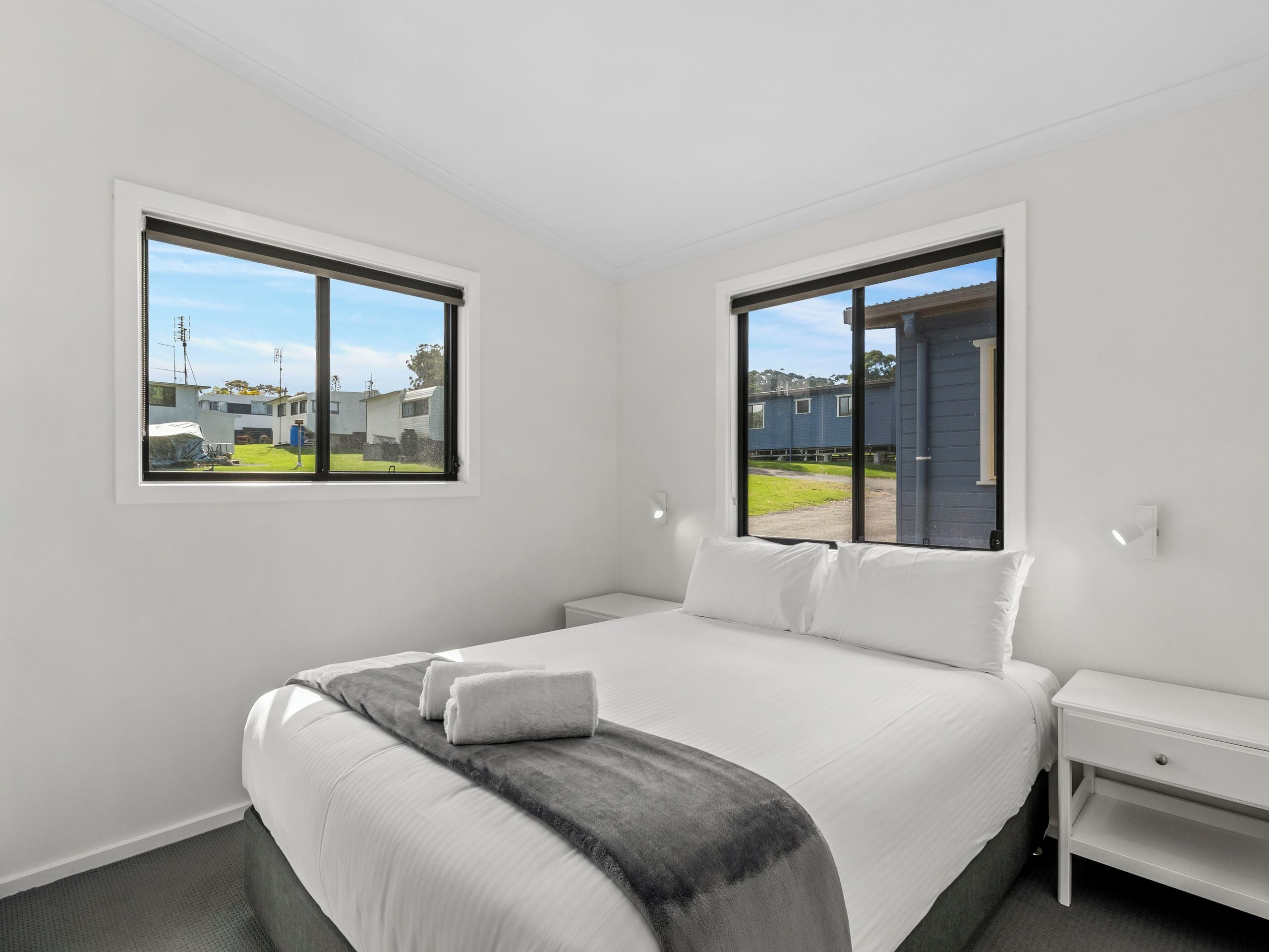 Une chambre moderne avec un grand lit, deux tables de chevet et une vue sur un quartier résidentiel, visible à travers deux fenêtres.