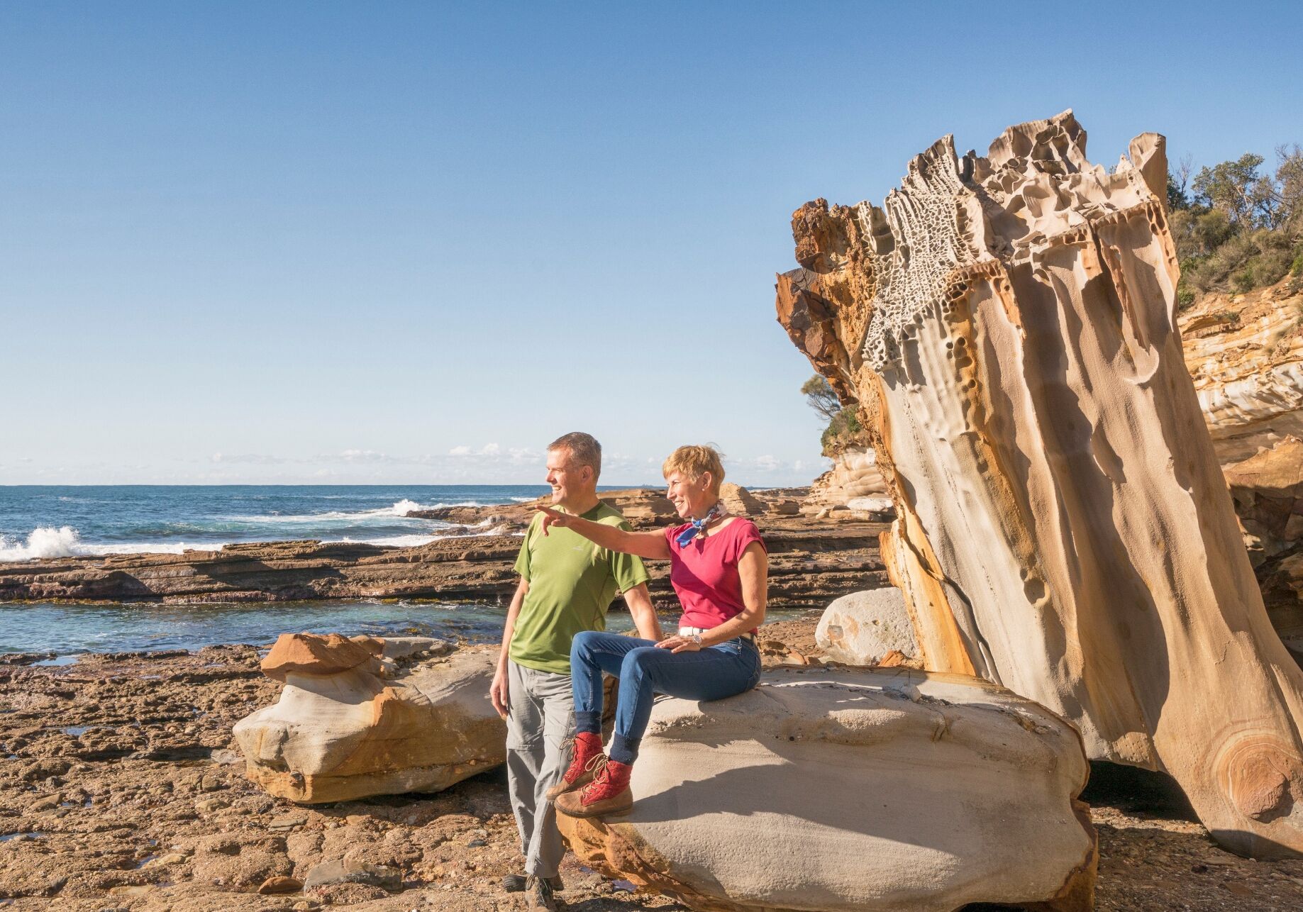 Deux personnes sont assises sur une plage rocheuse, avec une grande formation rocheuse en arrière-plan.
