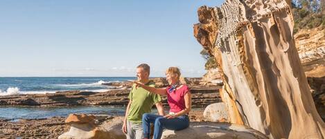 Two people sitting on a rocky beach with a large rock formation in the background.