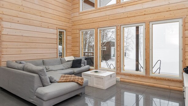 L’intérieur d’un chalet moderne en bois, avec un canapé gris, une table basse blanche et de grandes fenêtres donnant sur un paysage enneigé.