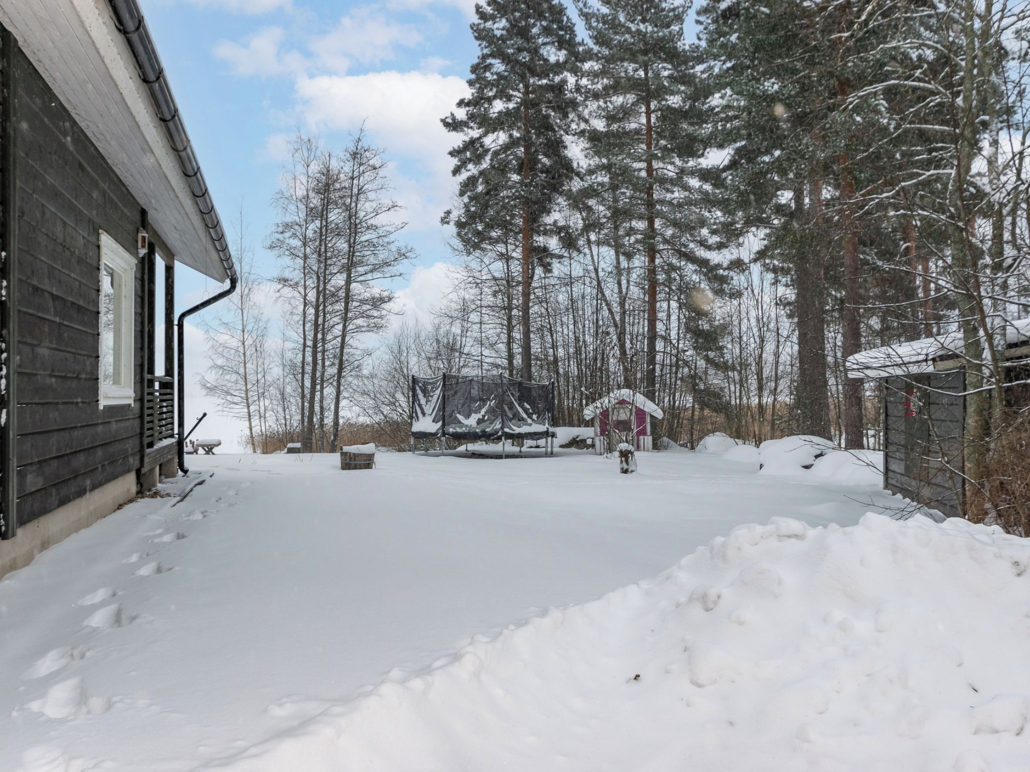 A snow-covered outdoor area with a wooden building, a covered structure, and tall trees.