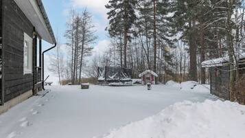 A snow-covered outdoor area with a wooden building, a covered structure, and tall trees.