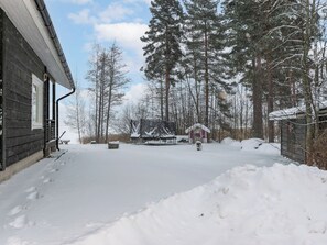 A snow-covered outdoor area with a wooden building, a covered structure, and tall trees.
