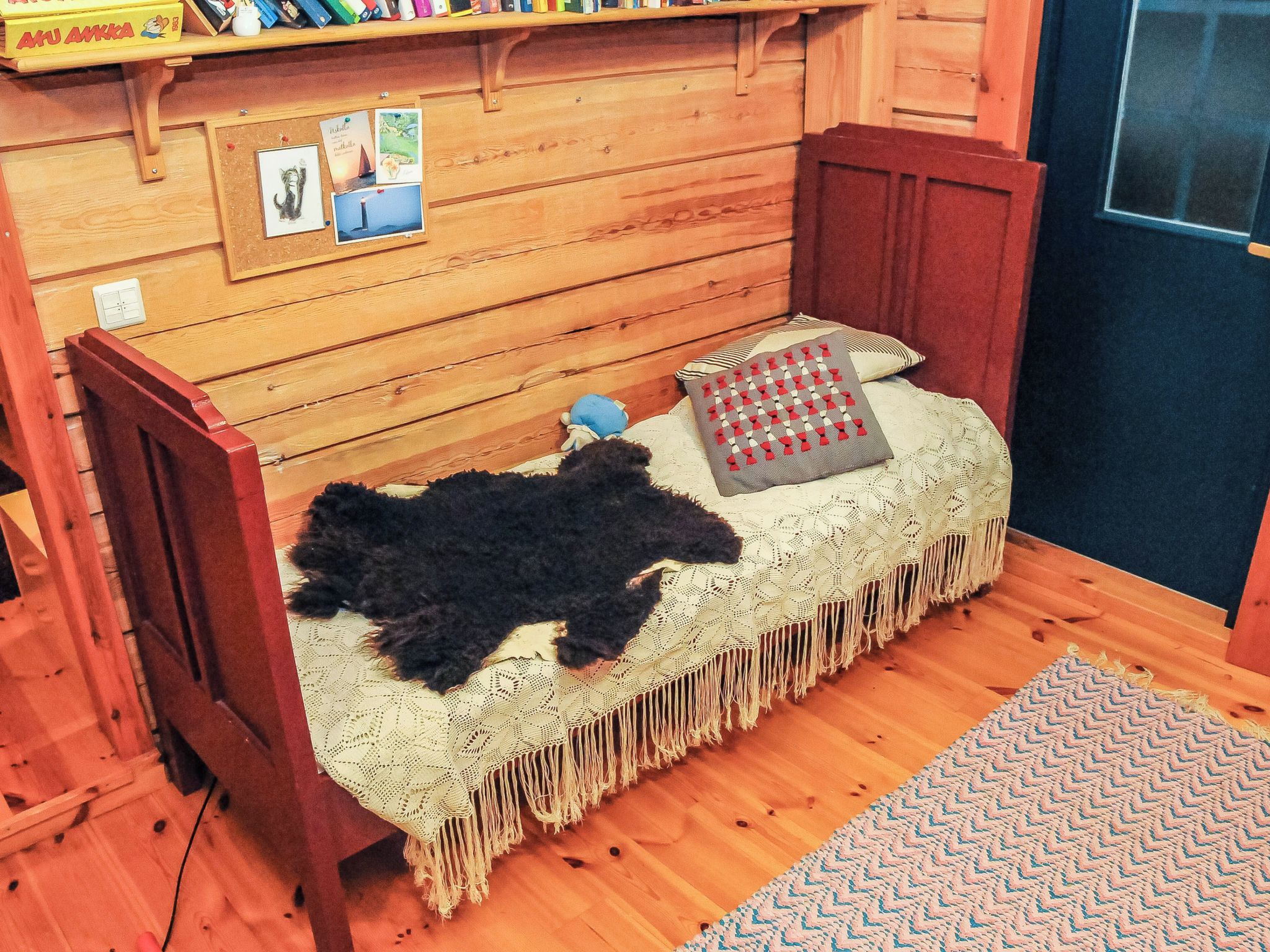 Ein gemütliches Zimmer mit einer Holzwand, einem kleinen Bett mit Strickdecke, einem roten Sessel und einem Regal mit Büchern und einer Kiste.