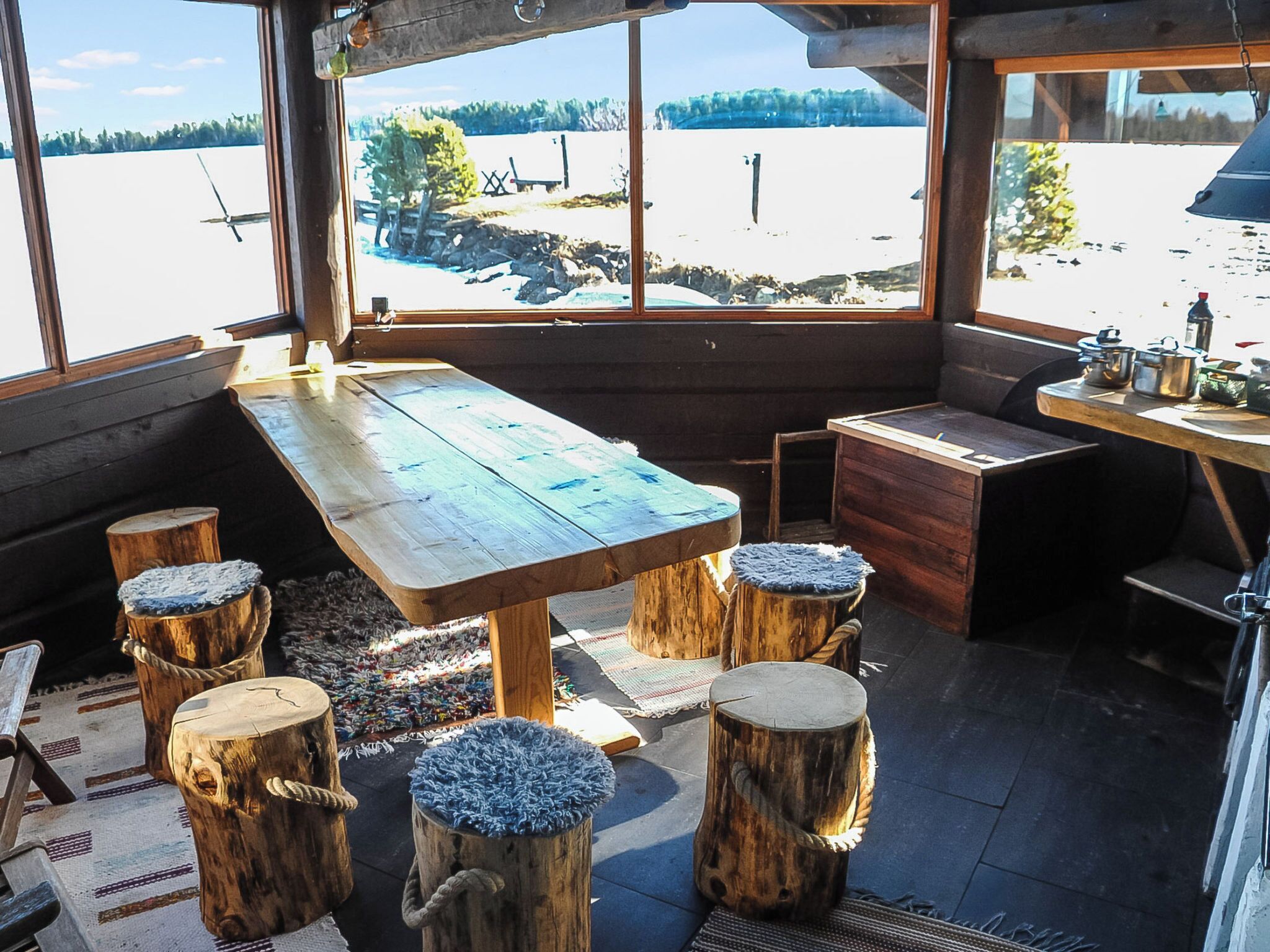 Une cabane en bois rustique, avec une grande table et des bancs, une vue sur un paysage enneigé par les fenêtres, et un petit coin cuisine équipé de casseroles et de poêles.