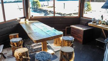 Une cabane en bois rustique, avec une grande table et des bancs, une vue sur un paysage enneigé par les fenêtres, et un petit coin cuisine équipé de casseroles et de poêles.