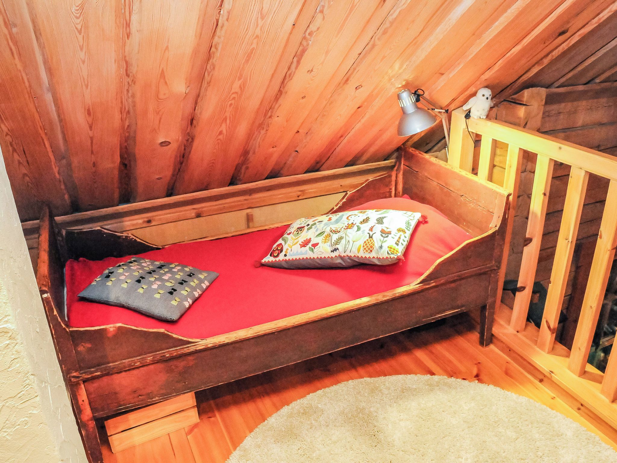 A small wooden bed with a red mattress, a pillow, and a lamp on a wooden nightstand.