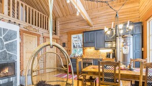 Un intérieur de chalet chaleureux avec une cheminée en pierre, un fauteuil suspendu, une table à manger en bois et un coin cuisine.