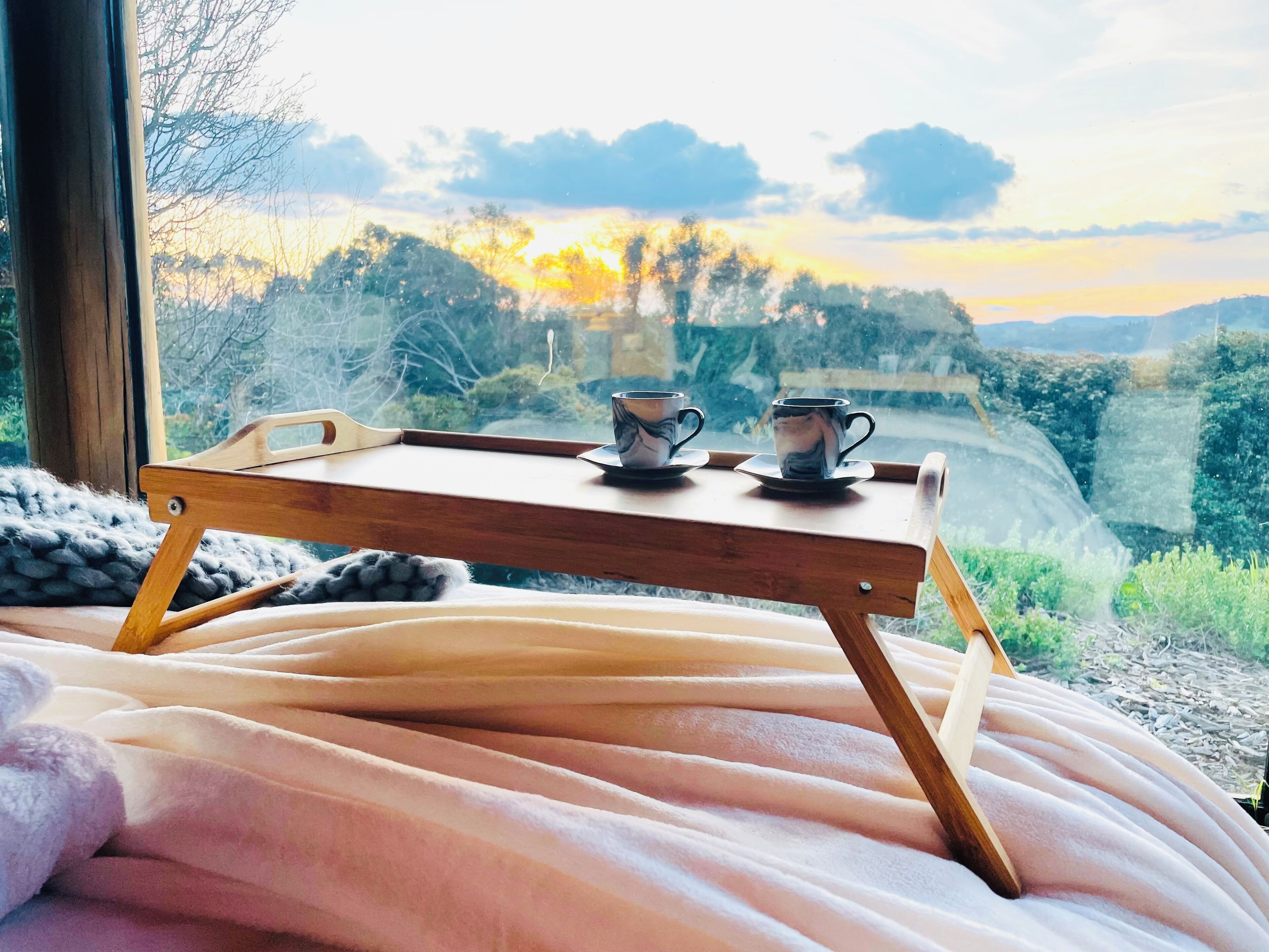 A bed with a wooden tray holding two cups, set against a window with a view of a landscape.