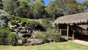 Una construcción con techo de paja y estructura de madera, rodeada por un jardín cuidado con rocas, arbustos y césped.