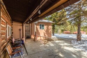 A covered outdoor seating area with a wooden table and chairs, a stone wall, and a view of a snow-covered landscape with trees and buildings in the background. - Medallion Cabin by Velo (Eagle Nest)