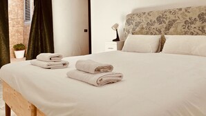 Un lit bien fait, avec des serviettes pliées, une tête de lit à motifs floraux et une lampe de chevet posée sur une table de chevet blanche.