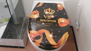 A toilet with a VIP invitation lid in a bathroom with white tiles and a glass shower enclosure.