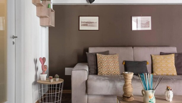 Een woonkamer met een grijze bank, twee ingelijste foto’s aan de muur, een klein rond tafeltje met een vaas en kaarsen, en een hoge vaas met een plant.