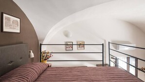 Een slaapkamer met een gebogen plafond, een bed met gestreepte lakens, een bedlampje en twee ingelijste foto's aan de muur.