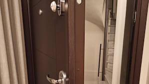 Une porte en bois, dotée d’une poignée moderne et d’un trou de serrure, qui donne sur un couloir au sol carrelé et où se trouve un escalier.