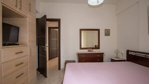 Une chambre à coucher comprenant un lit, une commode, un téléviseur posé sur un meuble, un miroir au-dessus de la commode et une porte donnant sur une autre pièce.