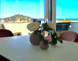 Ein Tisch mit drei Vasen voller Blumen, ein Fenster mit Blick auf einen Strand und einen Berg, und ein Stuhl im Vordergrund.