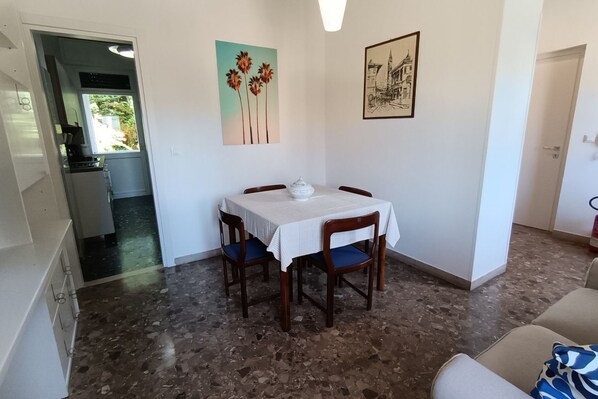 A dining area with a table, chairs, and a white tablecloth. There are two wall-mounted artworks, one with palm trees and one with a building. The room has a tiled floor and a door leading to another room.