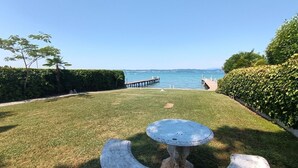 Un espace extérieur verdoyant avec une table et des chaises, une jetée en bois qui s’avance dans l’eau et une haie sur la droite.