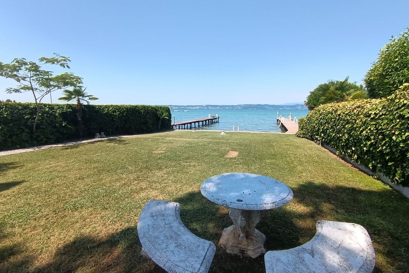 A grassy outdoor area with a table and chairs, a wooden dock extending into the water, and a hedge on the right.