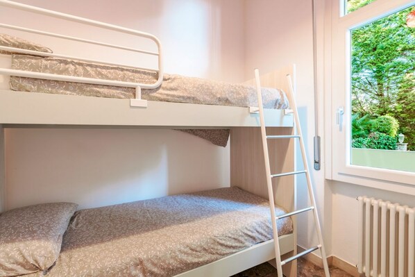 A bunk bed room with a ladder, a window with a view of greenery, and a radiator.
