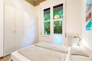 A bedroom with a bed, a window with a view of greenery, a radiator, and a wardrobe.