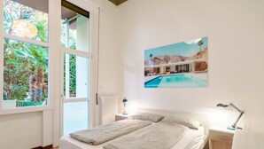 Un dormitorio con una cama, dos mesitas de noche, un ventanal con vistas a la vegetación y un cuadro de una piscina.