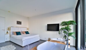 Ein modernes Schlafzimmer mit einem großen Bett, einer integrierten Badewanne, einem an der Wand befestigten Fernseher und einer Topfpflanze.