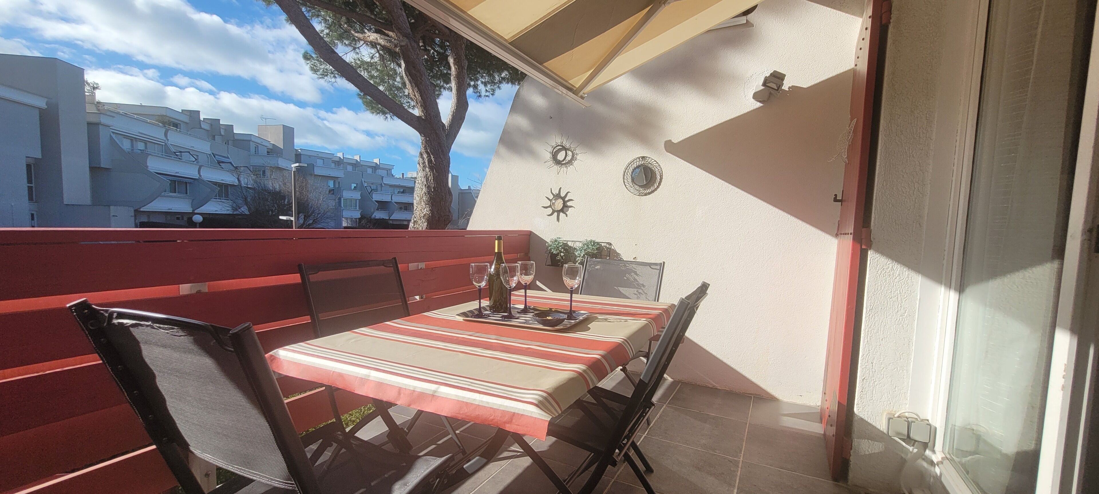 An outdoor dining area with a table set for two, a red bench, and a view of residential buildings.
