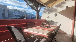 An outdoor dining area with a table set for two, a red bench, and a view of residential buildings. - Aux Portes du Golf, 5 mn de L'étang du Ponant, Proche de la Mer, Appartement T3 (La Grande-Motte)