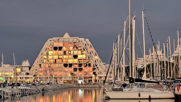 A marina with multiple boats docked, a large building with a unique geometric facade, and a calm water body reflecting the structure. - Aux Portes du Golf, 5 mn de L'étang du Ponant, Proche de la Mer, Appartement T3 (La Grande-Motte)