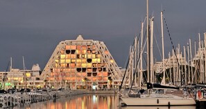 Un port de plaisance où sont amarrés de nombreux bateaux, un grand bâtiment doté d’une façade géométrique originale, et une étendue d’eau calme qui reflète la structure.