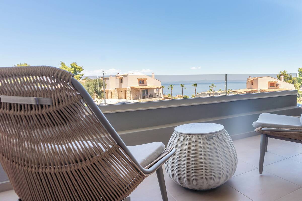 Une chaise et un pouf en osier sur un balcon offrant une vue sur les bâtiments et la mer.