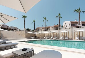 Zona de piscina con sillas reclinables y sombrillas, palmeras y un muro de piedra.