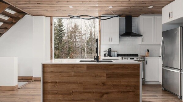 Een moderne keuken met een houten eiland, witte kasten, roestvrijstalen apparatuur en een groot raam met uitzicht op een bos.