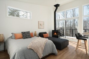 A modern bedroom with a large bed, a fireplace, and a view of trees through large windows. - The Mini-Méli - Romantic getaway in a cozy chalet (Sainte-Béatrix)
