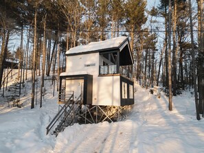 A cabin with a staircase leading up to a deck, surrounded by a snowy forest.