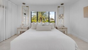 Een moderne slaapkamer met een groot bed, twee nachtkastjes en uitzicht op palmbomen door grote ramen.