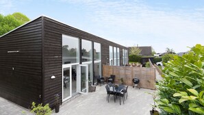 A modern house with a dark wooden exterior, large glass windows, and a patio area with outdoor furniture. - 2 bedroom gorgeous home in Gilleleje (Gilleleje)