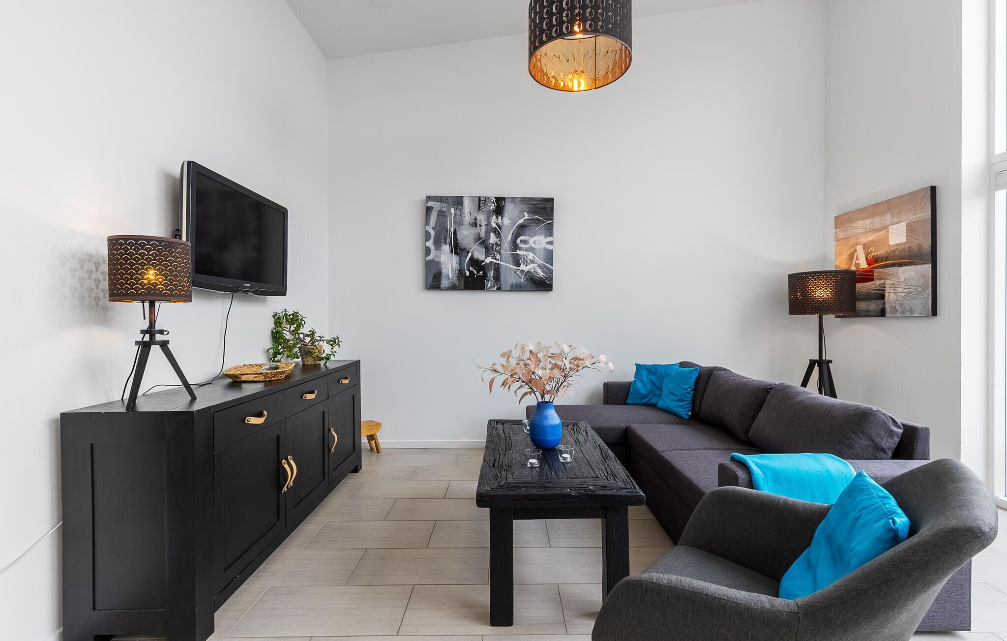 Een moderne woonkamer met een zwarte mediakast, een flatscreen televisie, een zwarte salontafel, een grijze bank met blauwe kussens en twee vloerlampen.
